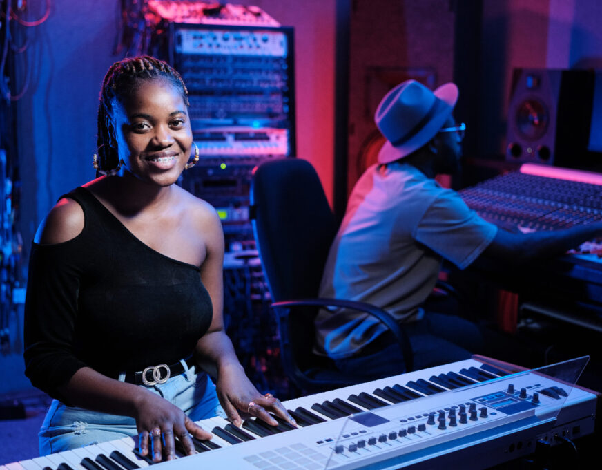 Cheerful Woman Working in Recording Studio Joyful young African American female musician playing digital keyboard while producer working on mixing console in recording studio