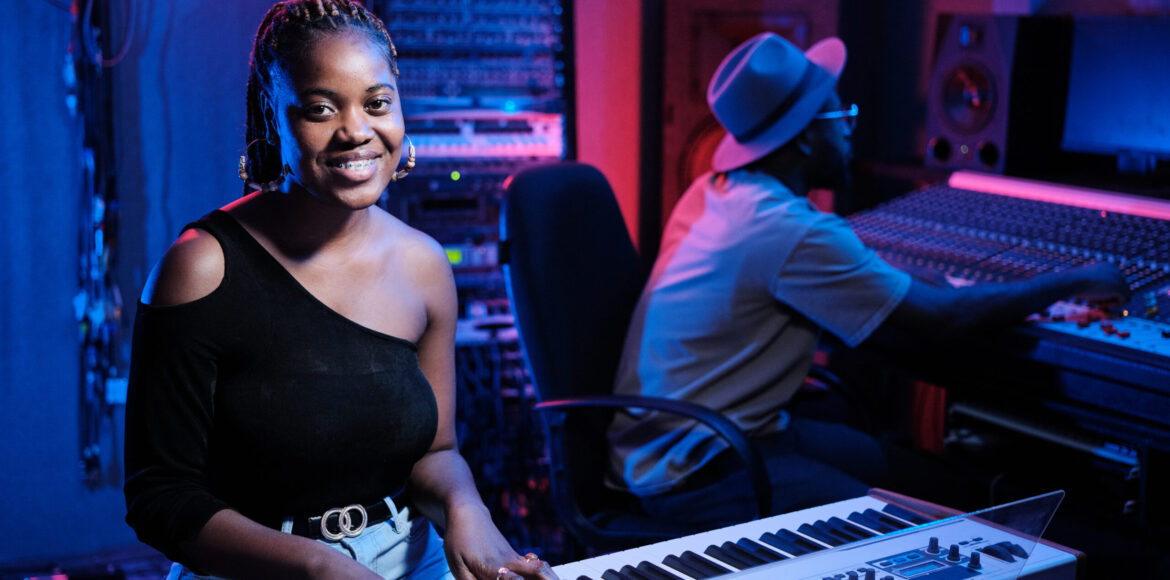 Cheerful Woman Working in Recording Studio Joyful young African American female musician playing digital keyboard while producer working on mixing console in recording studio