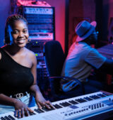 Joyful young African American female musician playing digital keyboard while producer working on mixing console in recording studio